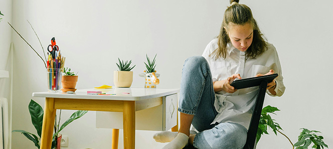 Girl in white shirt and jeans holding a tablet and sitting at a small table with plants.