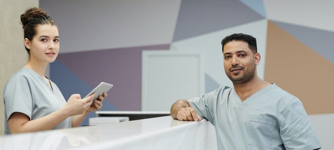 Woman standing behind a counter wearing scrubs, looking at the camera. Man standing on the opposite side, to the right, looking at the camera.
