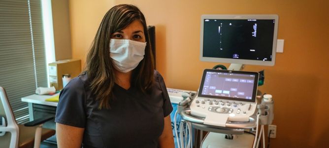 Woman with brown hair, wearing a surgical mask and gray scrubs, standing in front of medical equipment.