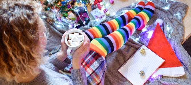 Young woman sitting on the floor  next to holiday gifts, holding a mug of cocoa with marshmallows and looking in a journal.